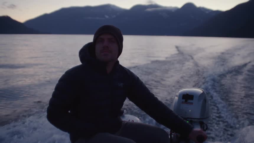 Man directs small motorboat through water as sky over misty mountains turns red
