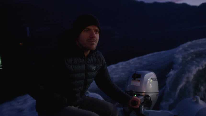 Man pilots small motorboat through water reflecting red sky with town lights in the distance