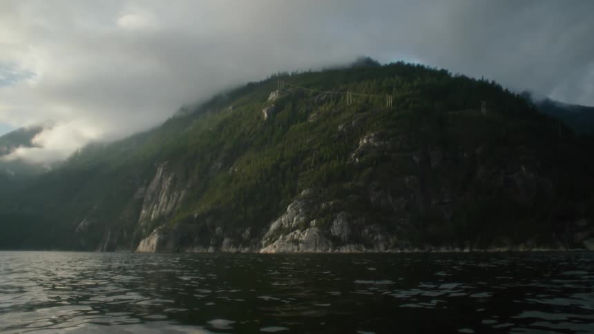 View from a boat of a forested mountain on the shore of an ocean on a cloudy afternoon