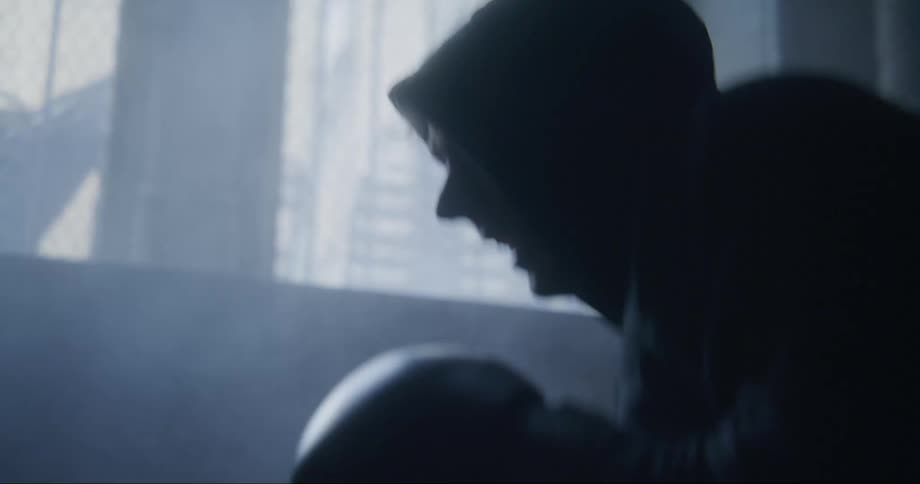 Young man wearing a hoodie and boxing gloves practices on a dimly lit room near a window