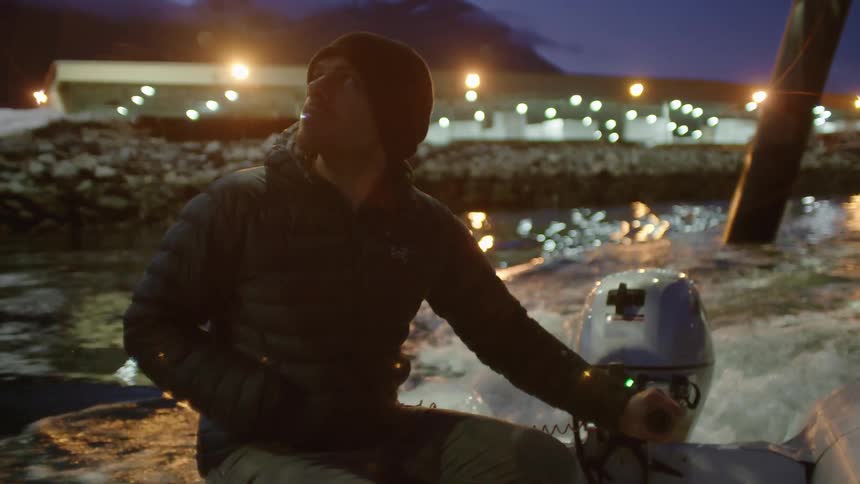 Man pilots small motorboat through water reflecting town lights at night