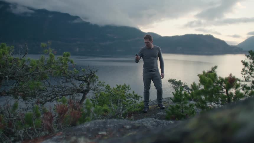A young man searching with a gps device steps onto a large boulder with a vast river and mountains behind him