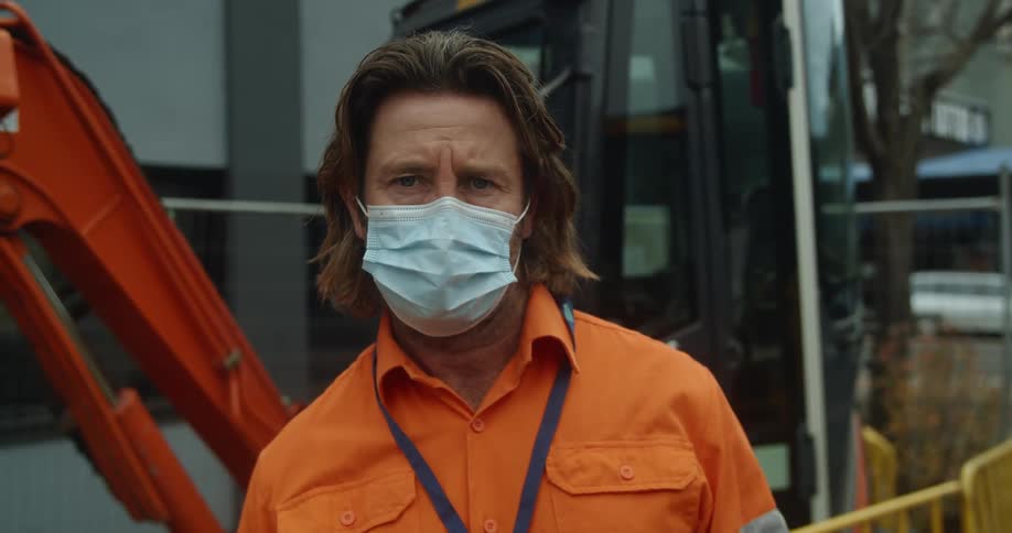 A man wearing an orange shirt and face mask stands outside next to a digger