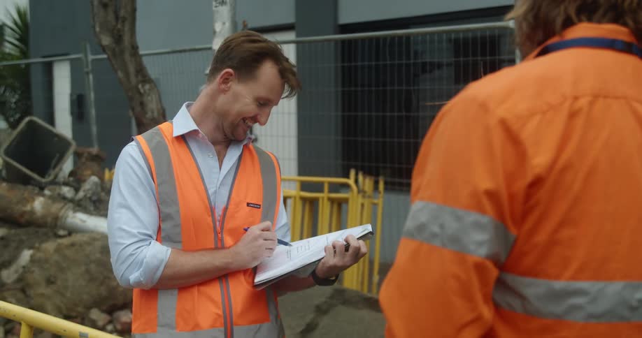 A man with a vest a pen and papers talking and smiling to another man