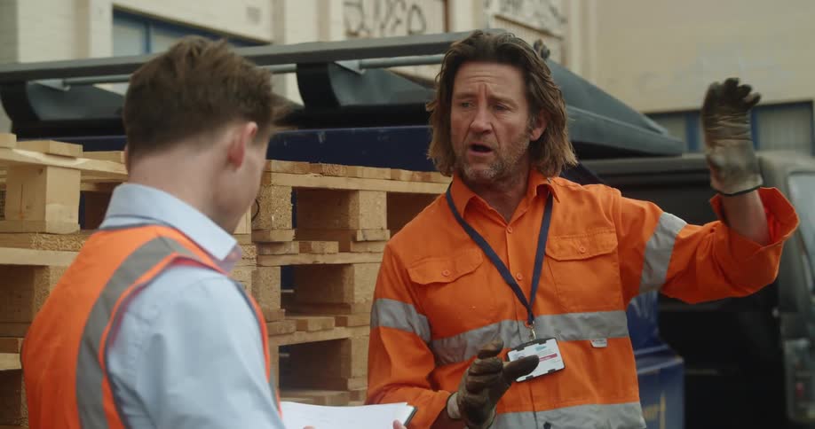 Two men wearing high visibility attire stand near stacked wooden palettes and van behind building discussing over notebook