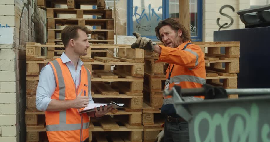 A pair of workers talk while standing outside of a building near a stack of wooden palettes