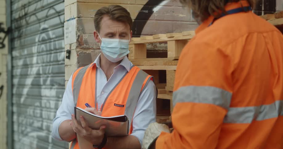 A man is taking notes as another coworker talks to him while standing outside next to a graffiti covered metal door with both men wearing orange safety tops