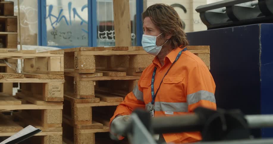 Man in orange reflective shirt is wearing a mask and work gloves while leaning on a pile of pallets talking using his hands to someone else who is holding a paper and a pen they are beside a few dumpsters and in front of a building with graffiti