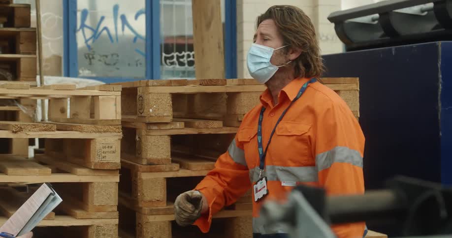 A man in a medical mask and reflective clothing talks with another person while leaning against a stack of wooden palettes outside of a building during the day