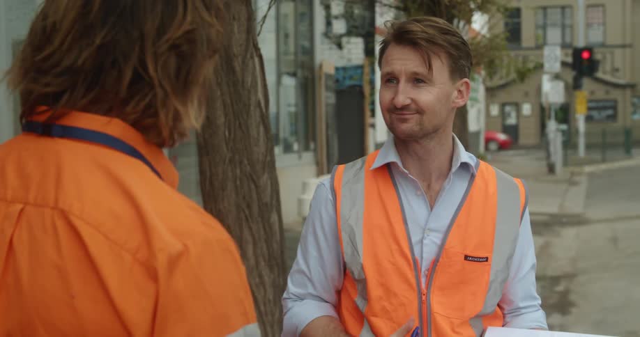A man with a vest a pen and a notebook standing in a city talking to another man