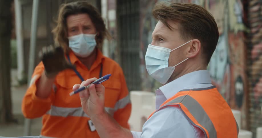 A man with a mask a vest a notebook and pen standing in a city and talking to another man