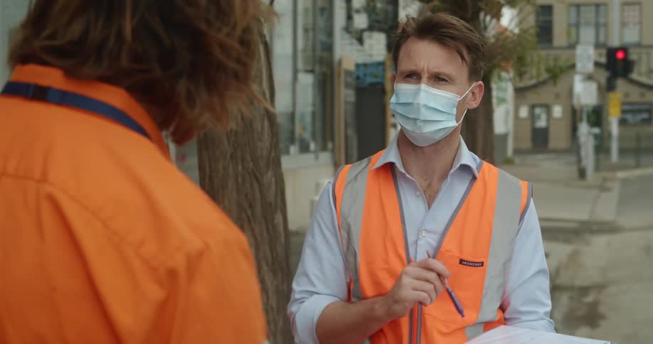A man with a mask a vest a pen and notebook standing in a city talking to another man