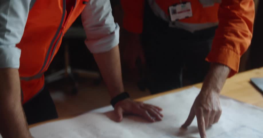 A man in an office marks a blueprint with a pen as another man standing beside him points and gestures at the blueprint