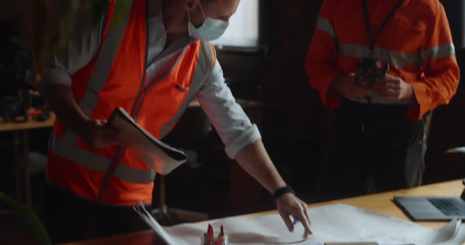 A pair of men in safety vests and medical masks examine a blueprint on a desk in an office during the day