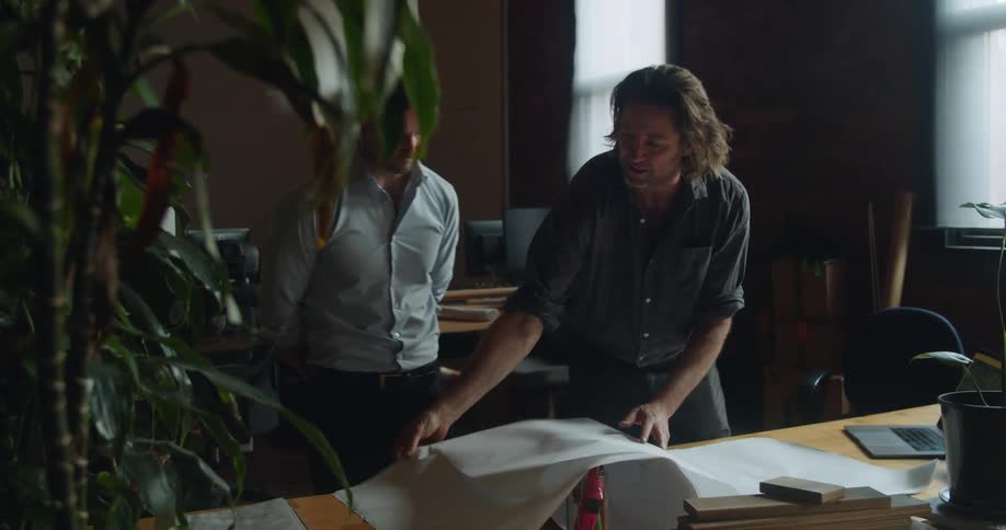 Men look at and discuss blueprints on table in office