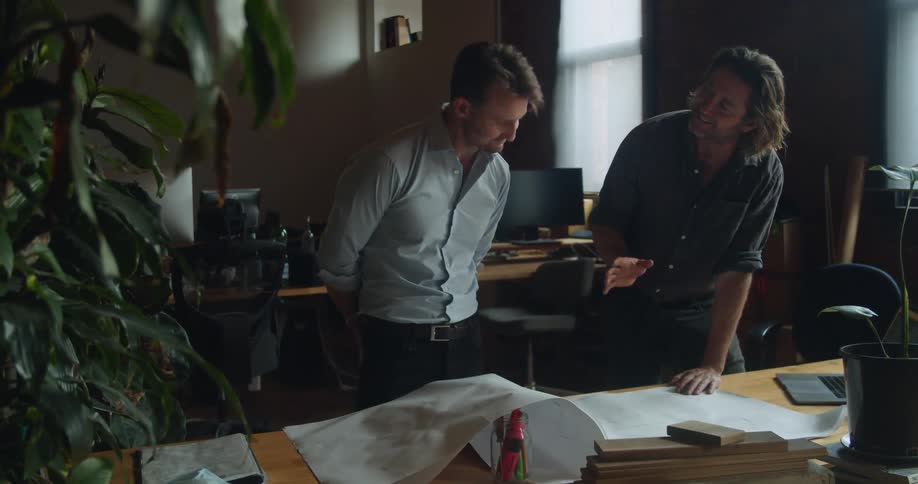 A pair of men in an office talk while looking at a blueprint
