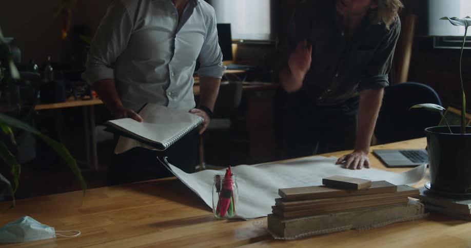 Two men in an office look at a blueprint and paperwork and talk