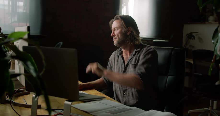 A man sitting at his desk raises his hand in greeting to the person he is having a video conference with