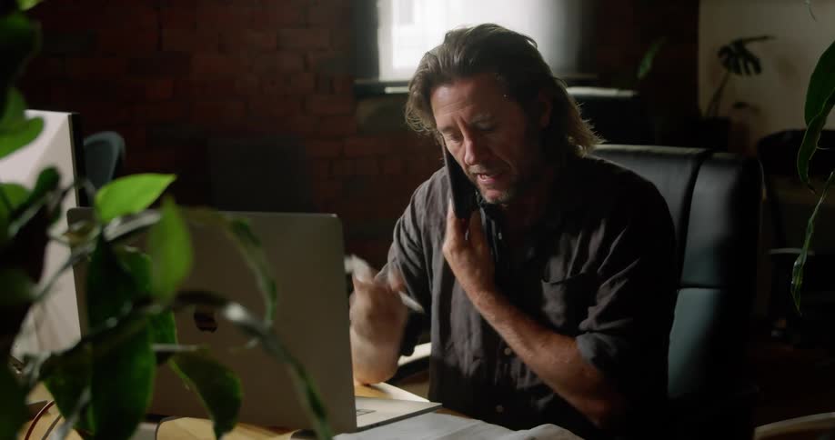 A man talks on the phone while sitting at his desk and writes something down
