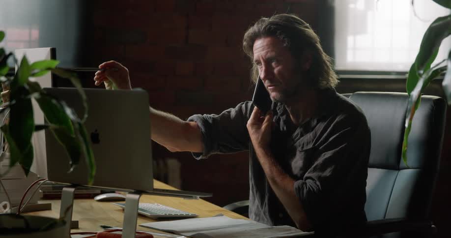 A man sits at his desk talking on the phone and gesturing at a monitor with the pen hes holding