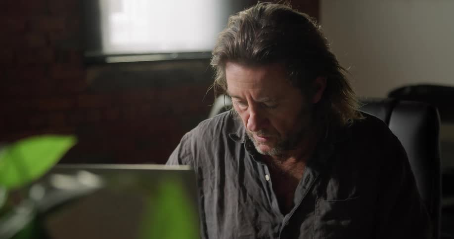 Long haired man sitting at the table uses a laptop in the office