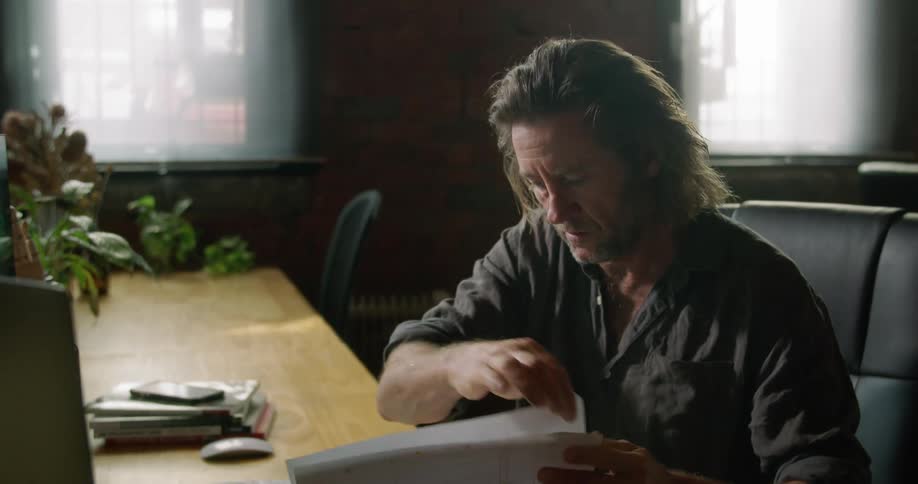 A man sits at a desk looking between a laptop and paperwork in front of him