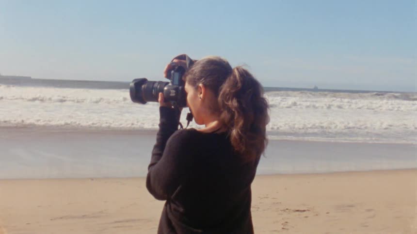 A photographer raises a camera to her face to photograph a beach during the day