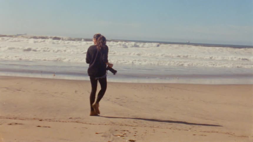 Photographer with wavy hair glasses black sweatshirt and pants walks down the beach near the ocean