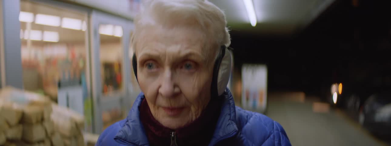 An older women wearing a blue jacket stands drinking out of a straw and then starts walking