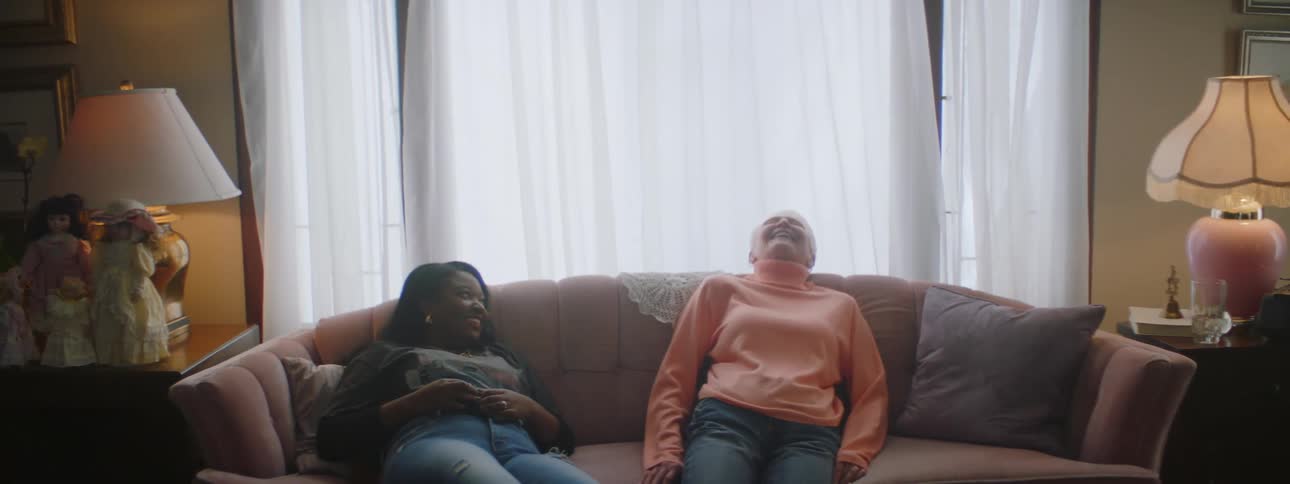 An elderly woman walks across the living room and sits on the couch with another woman and they laugh