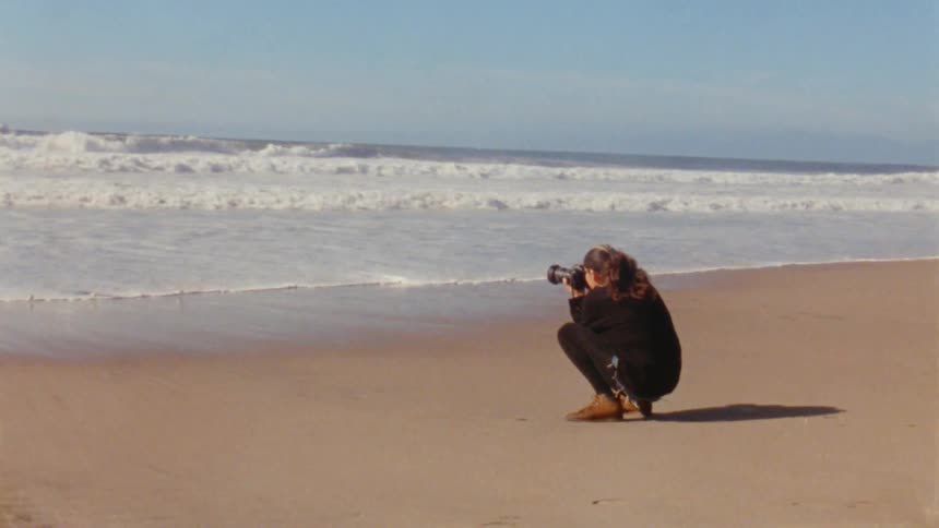 A woman is squatting down on the beach as she takes pictures of the waves rolling into shore