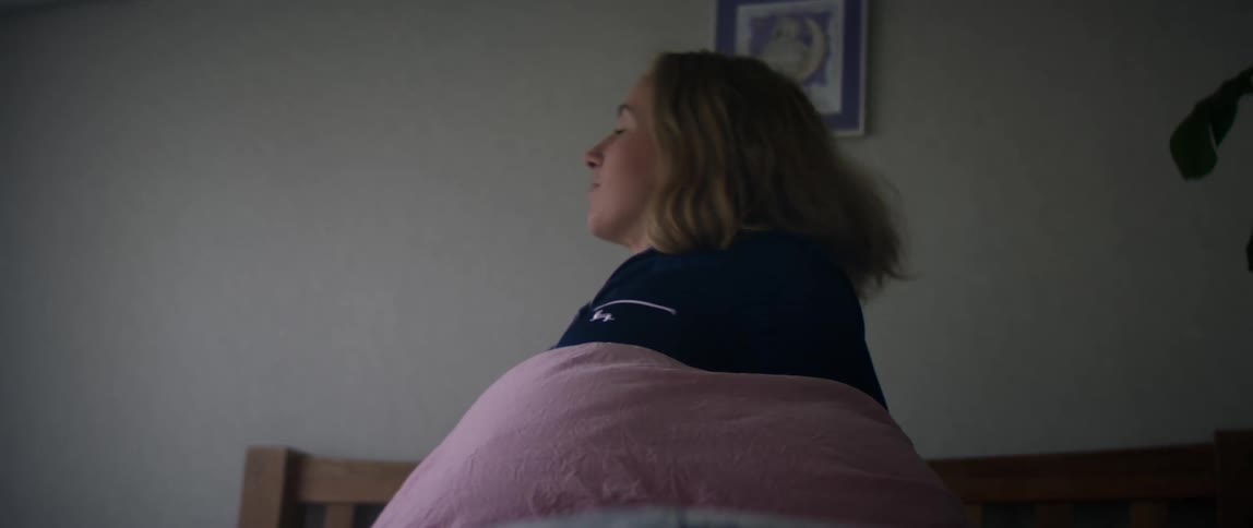 A woman sits up in bed and moves her blanket as she looks around