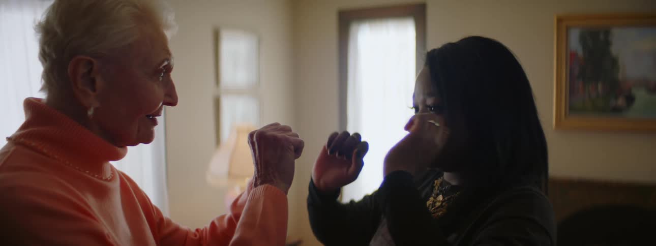 An elderly woman is standing in the living room with a younger woman as they are doing tai chi movements together
