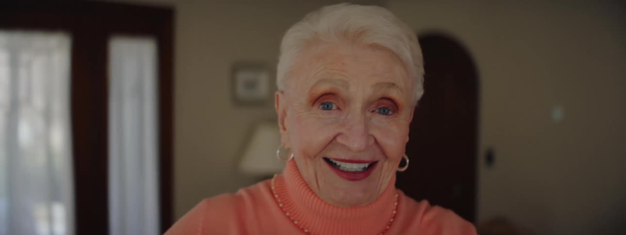 An elderly woman is smiling and laughing while turning around in the living room while looking at the camera