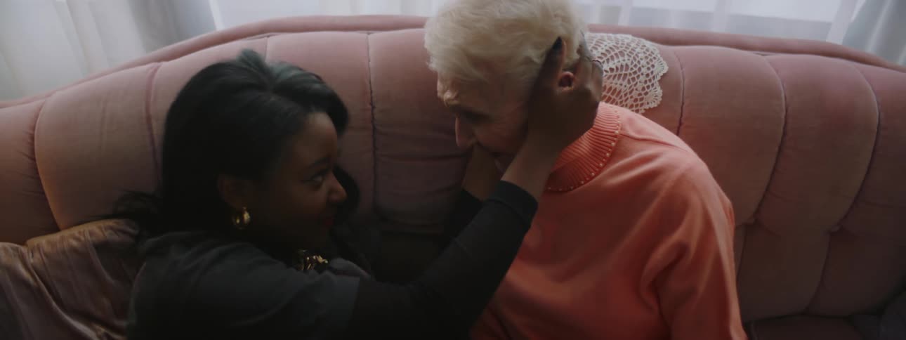 An elderly woman at home sits on her couch with a young black woman and they laugh and then put their heads together
