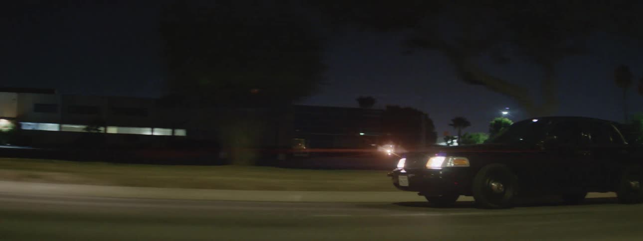 The headlights of a black automobile shine as it drives by buildings and parking lots at night