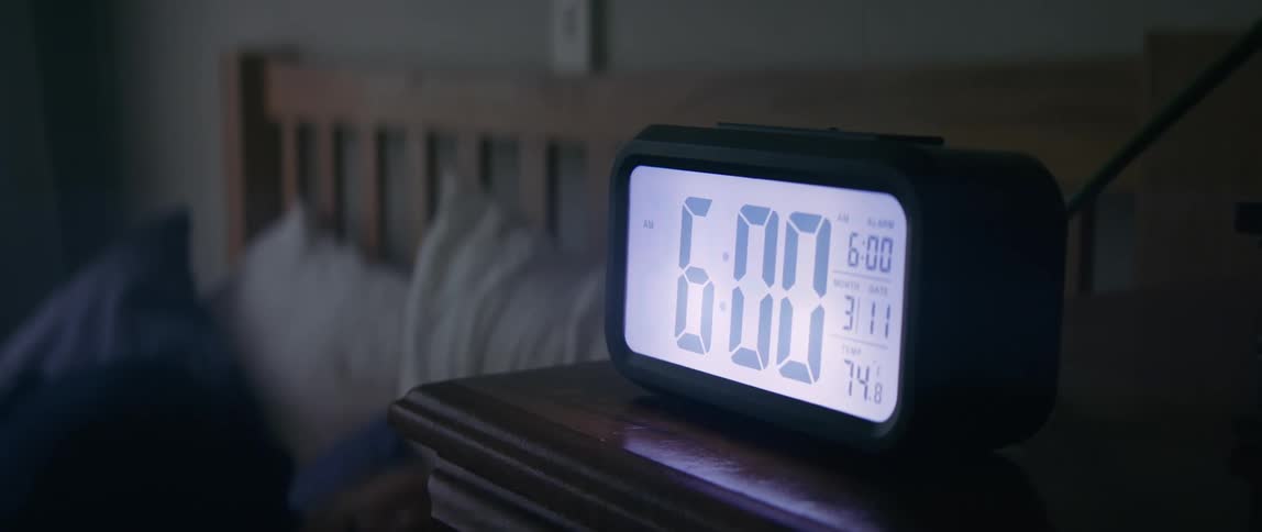 A man hitting a snooze button on an alarm clock in the morning