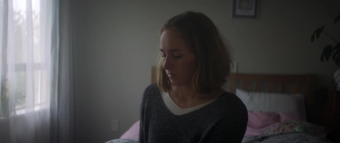 Woman with brown hair and gray sweater sits on bed using smartphone