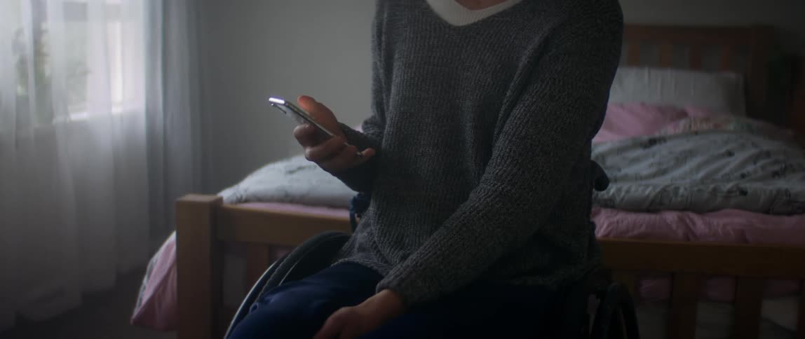 A women sitting in a wheelchair in front of her bed using her cell phone
