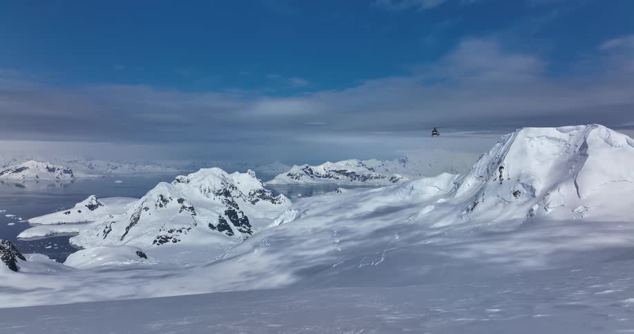 A helicopter is flying over snow covered mountains under a blue cloudy sky