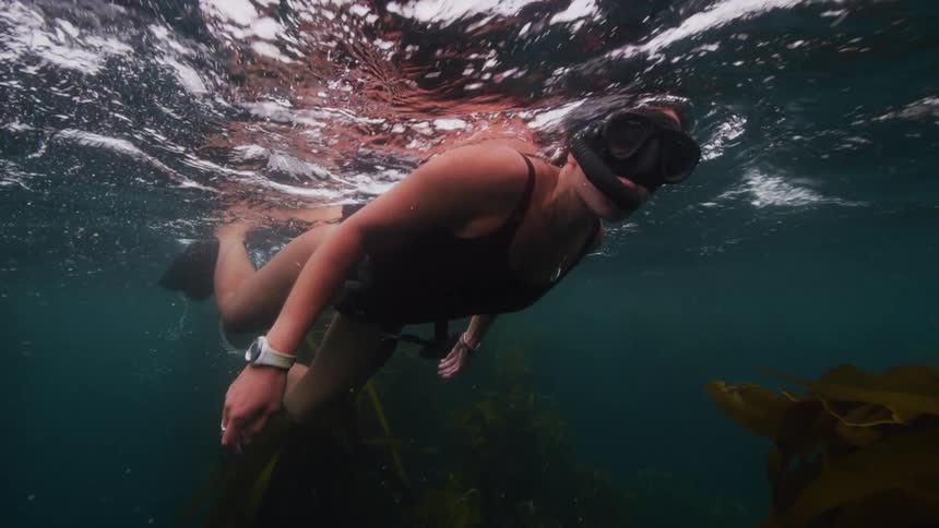 A woman swimming underwater with goggles and a snorkel among seaweed