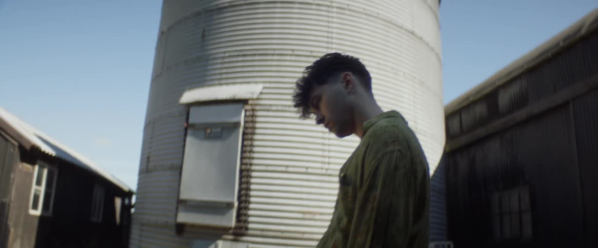 A sad looking teenage boy stands next to a silo and looks up in to the sky