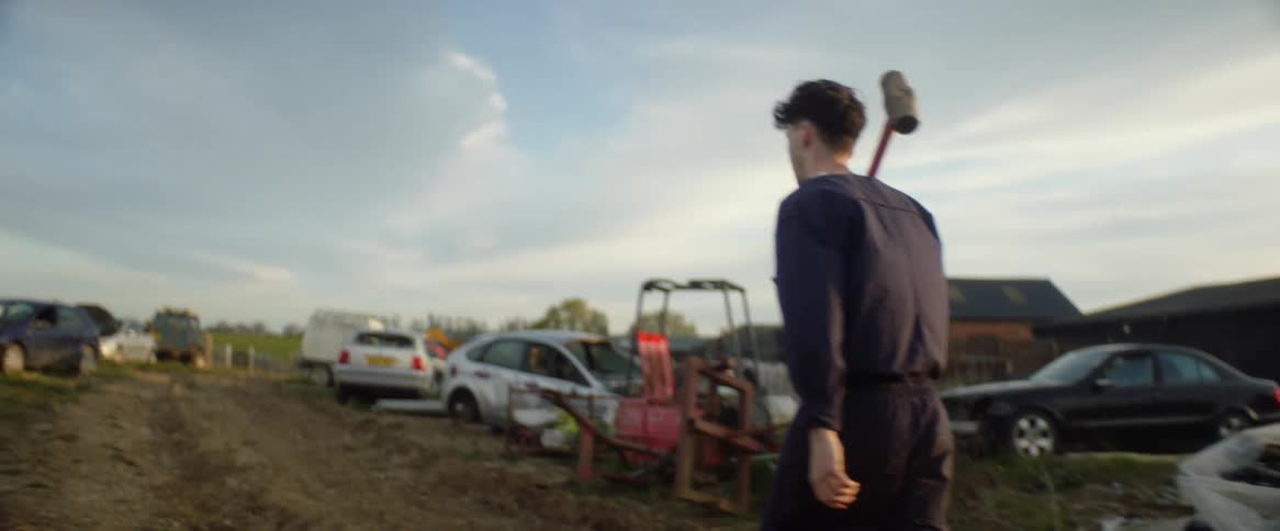 A man carries a heavy mallet through a junk yard filled with cars