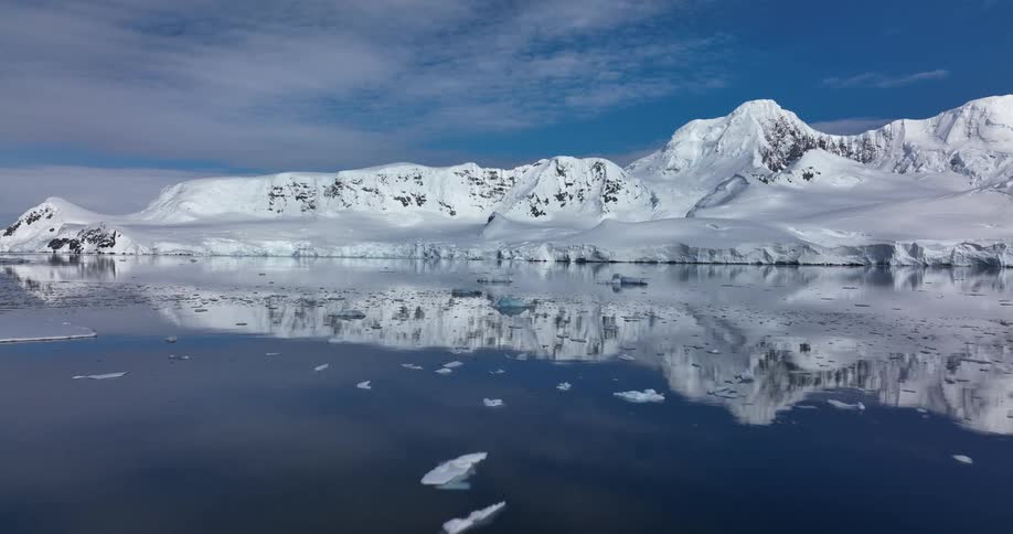 Pieces of ice float across the water with snow covered mountains along the coast as they reflect on the water surface