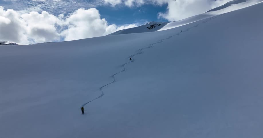 Three or four snowboarders riding down a snow covered mountain with blue skies and clouds in the background