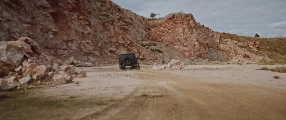Jeep kicks up dust as it drives along dirt road near rocky hill