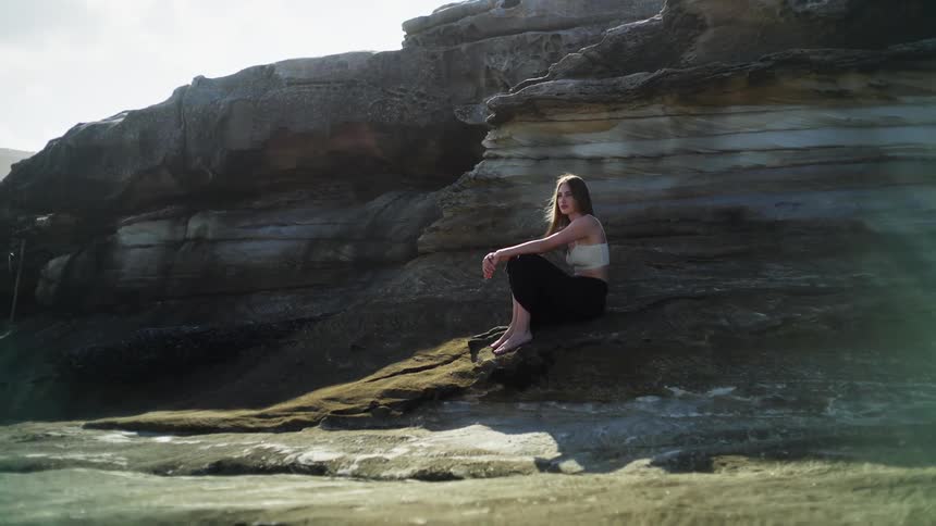 The sun shines on a woman with long dark hair sitting barefoot on a rock slope at the foot of a mountain