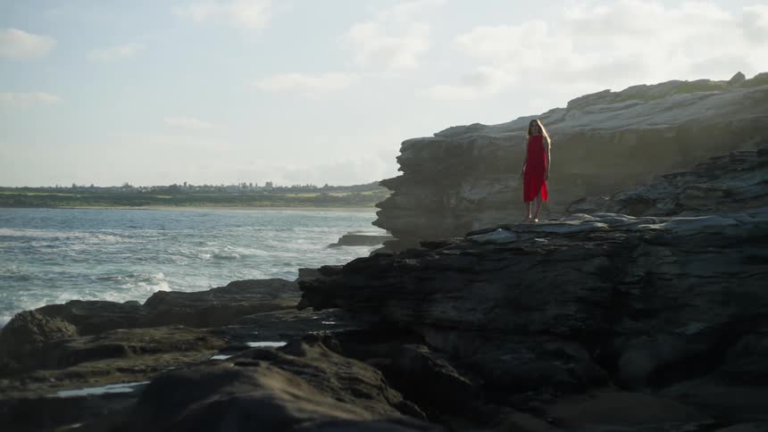 A woman in a red dress stands on the edge of a coastline cliff with waves crashing