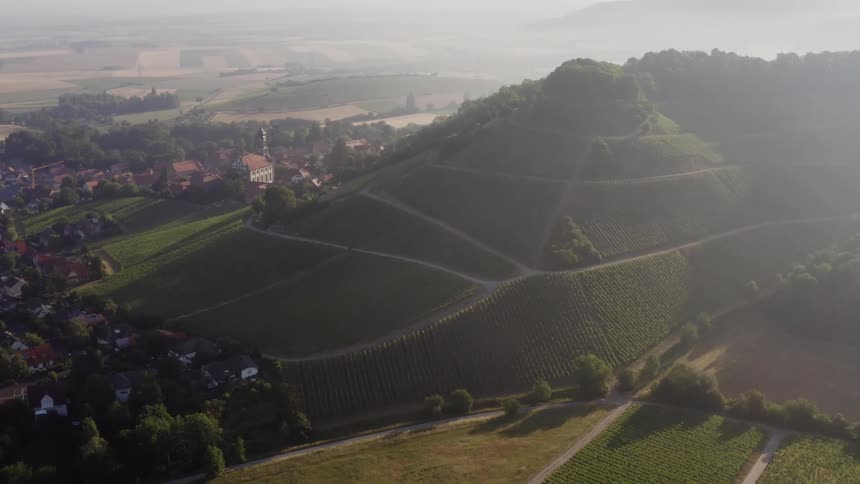 An aerial orbiting shot of a green hillside with lush fields and a town at the bottom of the hill