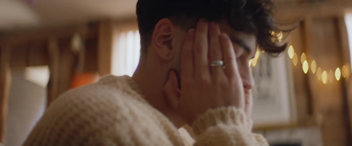 A man wearing a sweater rubs his hands together then massages his temples in contemplation while sitting indoors